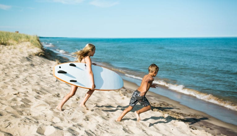 Two young children running toward Lake Michigan with a surfboard at Weko Beach.