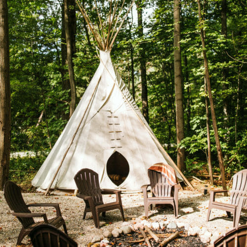 A circle of Adirondack chairs surround a firepit with a teepee and the woods as backdrop at Flamingo Ranch in Lakeside, Michigan.