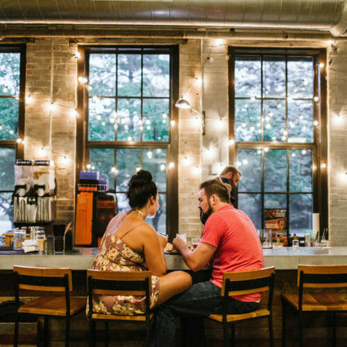 A couple sitting at the bar backed by large industrial windows at Journeyman Distillery in Three Oaks, Michigan.