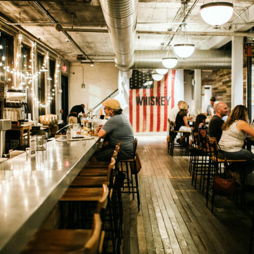 String lights reflecting off the long bar and a large American flag with the word 'whiskey' embroidered on it at Journeyman Distillery in Three Oaks, Michigan.