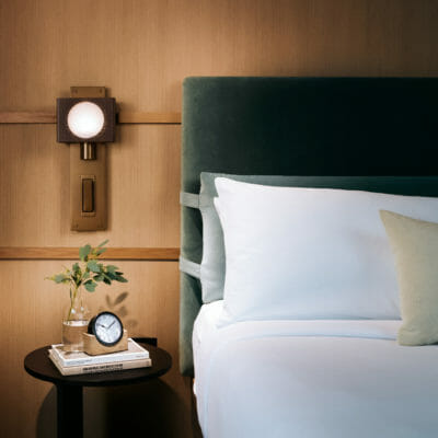 Bed with green velvet headboard and crisp white linens, with unique wall sconce and wood paneled wall in a guestroom at The Harbor Grand Hotel in New Buffalo, Michigan.