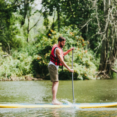 A bearded young man in an orange life vest and shorts paddling on a stand-up board on Galien River Marsh Train in New Buffalo, Michigan.