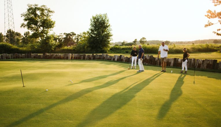 Two couples on a putting green with a rustic wood fence and open farm fields in the background in Three Oaks, Michigan.