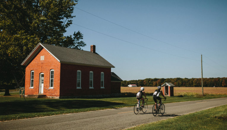 Two cyclists driving past an old brick schoolhouse on a country road during Apple Cider Century bike tour.