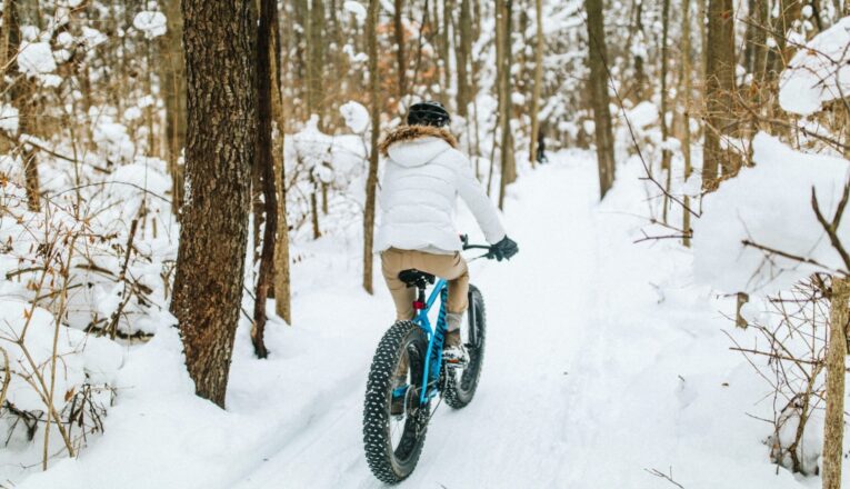 A woman in a white parka and tan pants riding a blue fat tire bike on a snowy wooded trail.