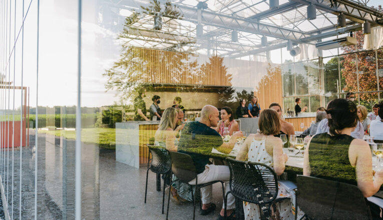A view through greenhouse windows of people smiling and conversing at long dining tables at Granor Farm in Three Oaks, Michigan.