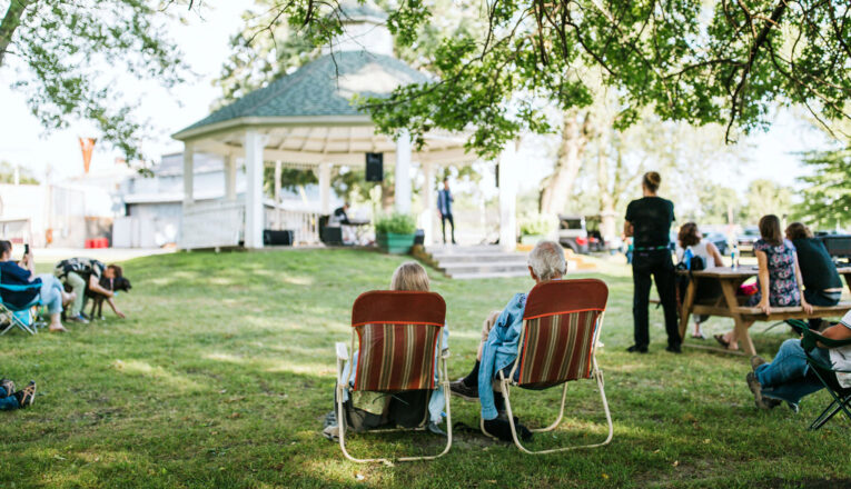 A couple sits in lawn chairs listening to music surrounded by mature oaks, green grass, and a vintage gazebo at Music in the Park in Three Oaks, Michigan.