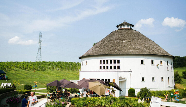 A white, round barn exterior with a large group of people sitting outside tasting wine in Baroda, Michigan.