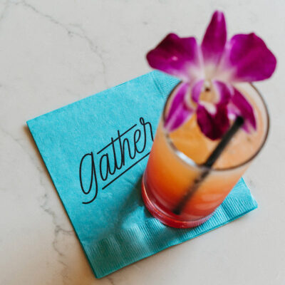 A colorful cocktail with a flower garnish sitting on a bright blue napkin at Gather in Harbert, Michigan.