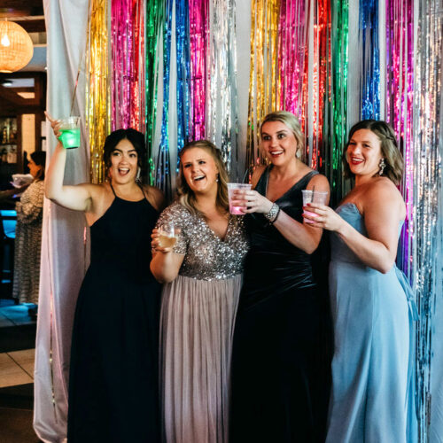 Four women pose for a prom-style photo in front of colorful streamers at the Harbor Country Pride Prom event in Union Pier, Michigan.
