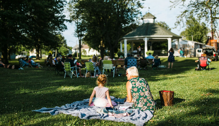A mother and daughter sit on a blanket listening to music surrounded by mature oaks, green grass, and a vintage gazebo at Music in the Park in Three Oaks, Michigan.