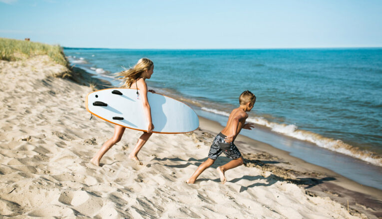 Two young children running toward Lake Michigan with a surfboard at Weko Beach.