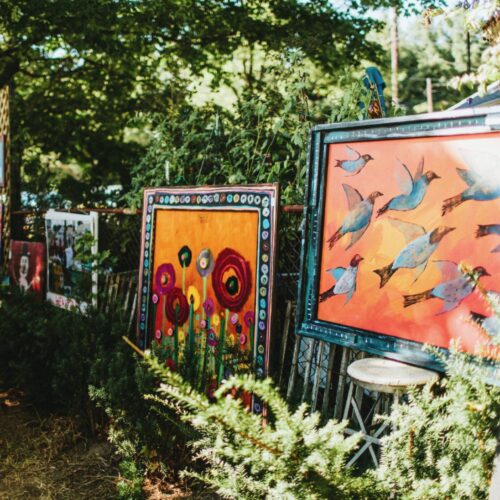A collection of large paintings displayed along a fence at Outsiders Outside Art