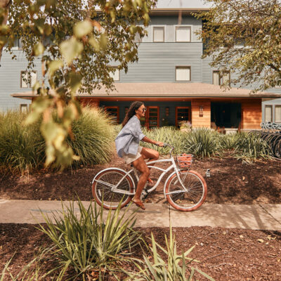 A woman joyfully bikes on a tree-lined path past the entrance of Harbor Grand Resort in New Buffalo, Michigan.