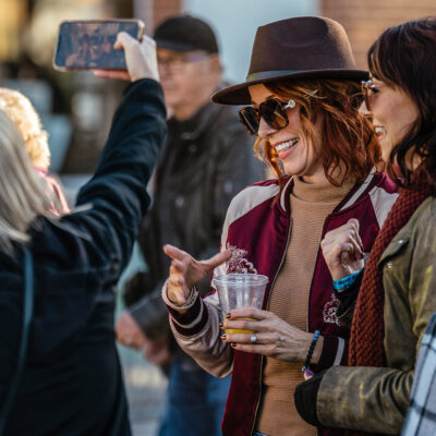 Women in sweaters, jackets, and hats smiling and taking selfies at New Buffalo Harvest and Wine Festival.