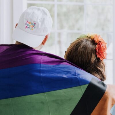 Friends wrapped in a colorful Pride flag gaze out a window.