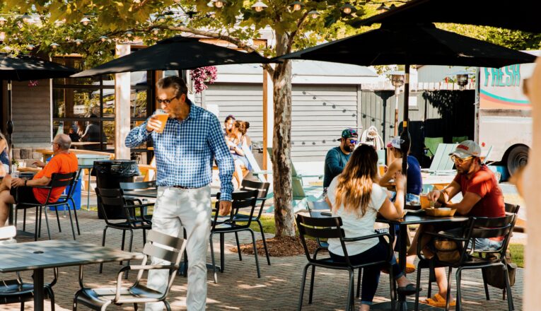 People in t-shirts, caps, and sunglasses chatting over drinks and food-truck fare on the patio at Sideyard in Sawyer, Michigan.