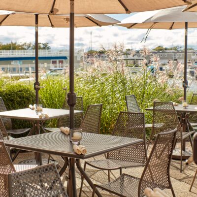 Dining tables with umbrellas surrounded by tall grasses and New Buffalo harbor on the patio at Terrace Room in New Buffalo, Michigan.