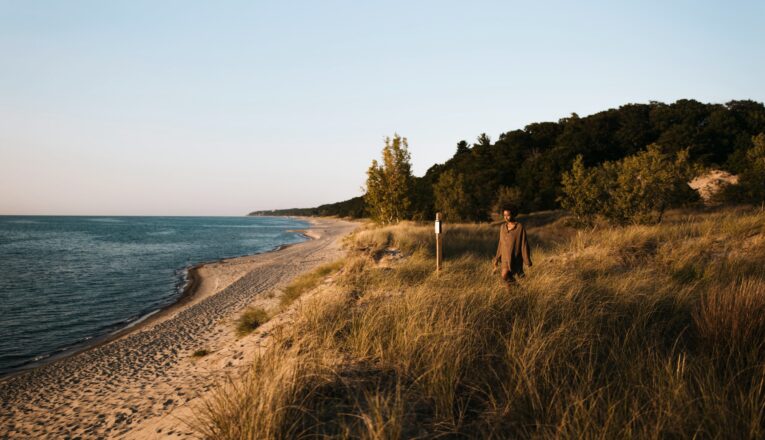 Young woman walking along a grassy dune trail toward a long, quiet expanse of Lake Michigan beach at Warren Dunes State Park.