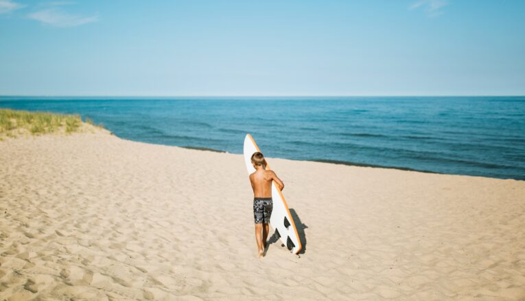 Young boy carrying a surfboard across a wide expanse of sand toward Lake Michigan at Weko Beach in Bridgman, Michigan.