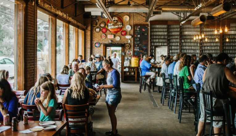 A rustic brick and wood taproom with vintage beer signs and Edison lights filled with people drinking and eating.