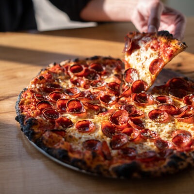 A close-up of a fresh pepperoni pizza on a wood table with a hand picking up a slice at Emma Hearth & Market in Bridgman, Michigan.