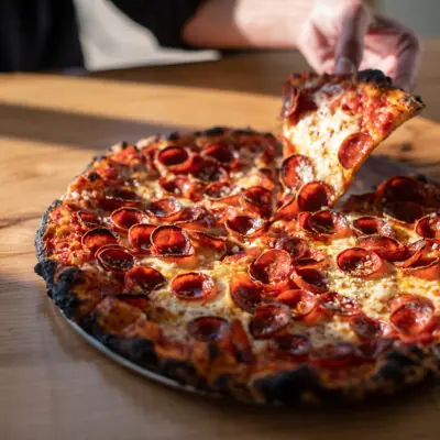 A close-up of a fresh pepperoni pizza on a wood table with a hand picking up a slice at Emma Hearth & Market in Bridgman, Michigan.