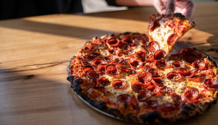 A close-up of a fresh pepperoni pizza on a wood table with a hand picking up a slice at Emma Hearth & Market in Bridgman, Michigan.