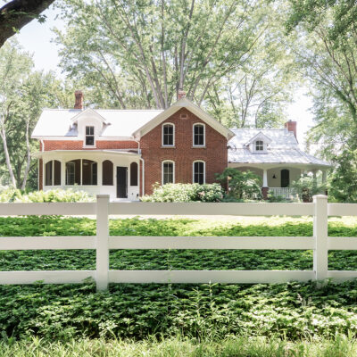 A charming old brick home sits back behind a white fence amongst old growth trees and lush groundcover.
