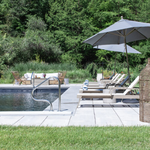 Sunchairs, umbrellas, and an outdoor seating area overlook a deep blue pool surrounded by a forest.