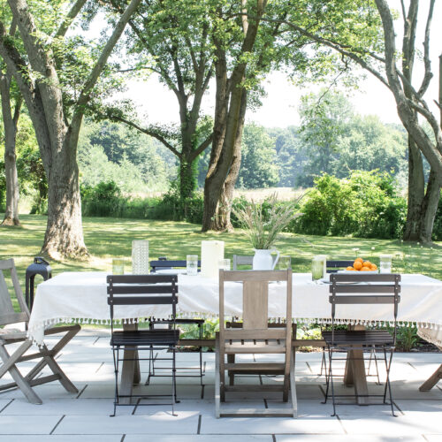 An outdoor patio with a white tablecloth-covered dining table with fresh fruit surrounded by old-growth trees.