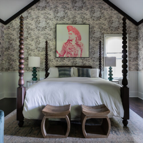 A bedroom with a four-poster bed, floral wallpaper, high ceilings, and a portrait in red above the bed.