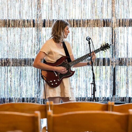 A woman tunes a guitar in front of empty wood chairs and metallic streamers in the window in the background.