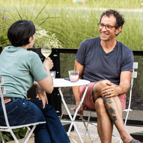A woman and a man sit outside at a white metal table with beverages enjoying a conversation.