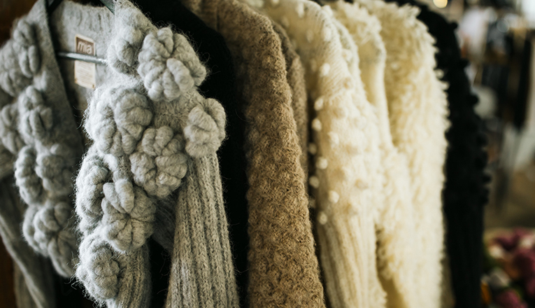 Heavy knit sweaters in neutral colors hang on a rack inside Goods and Heroes in Three Oaks, Michigan.