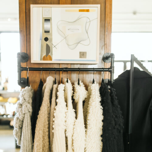 Framed artwork is displayed above a rack of neutral knits inside Goods and Heroes in Three Oaks, Michigan