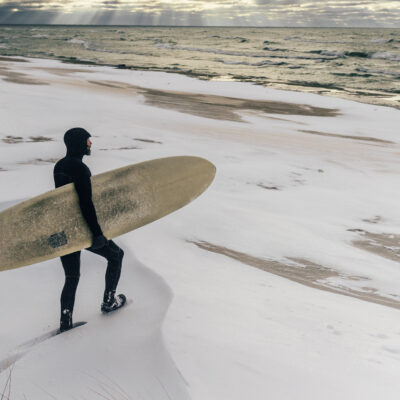 A surfer wearing a hoods, gloves, and booties walking through snow to go surfing in Lake Michigan in winter