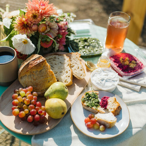 An array of food including freshly baked sourdough, produce, cheese, dips, and flowers atop a cafe table outside at Farmette in New Buffalo, Michigan